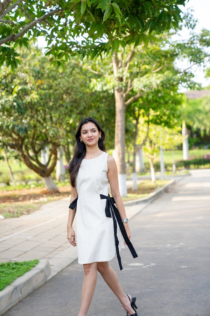 A woman wearing a sleeveless white knee-length dress with a black waist tie walks along a paved path in a park with green trees.